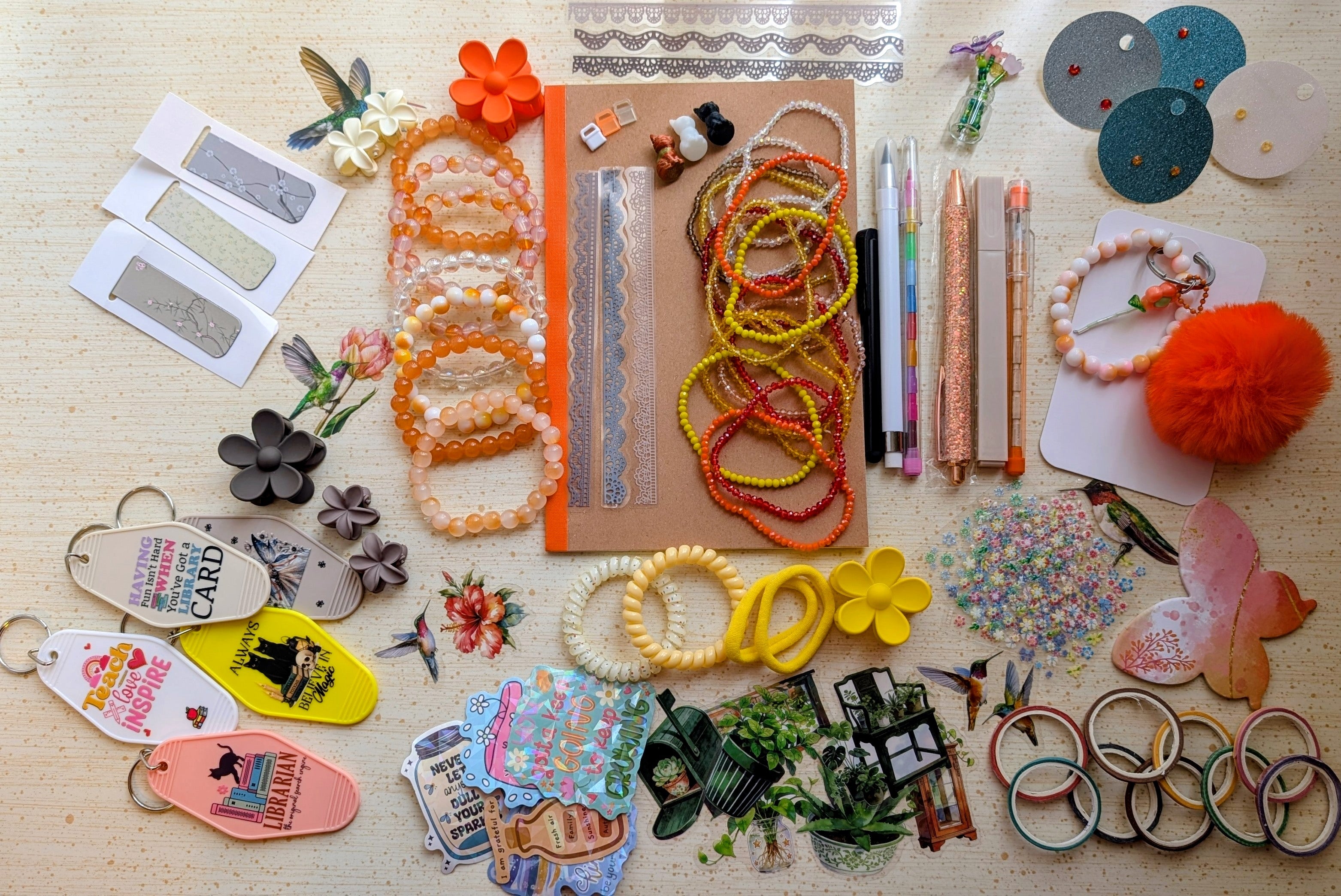 A flat-lay display of various handmade beaded bracelets, keychains, magnetic bookmarks, stickers, stationary items suck as pens, pencils, a notebook, and small craft accessories arranged on a light wood surface.
