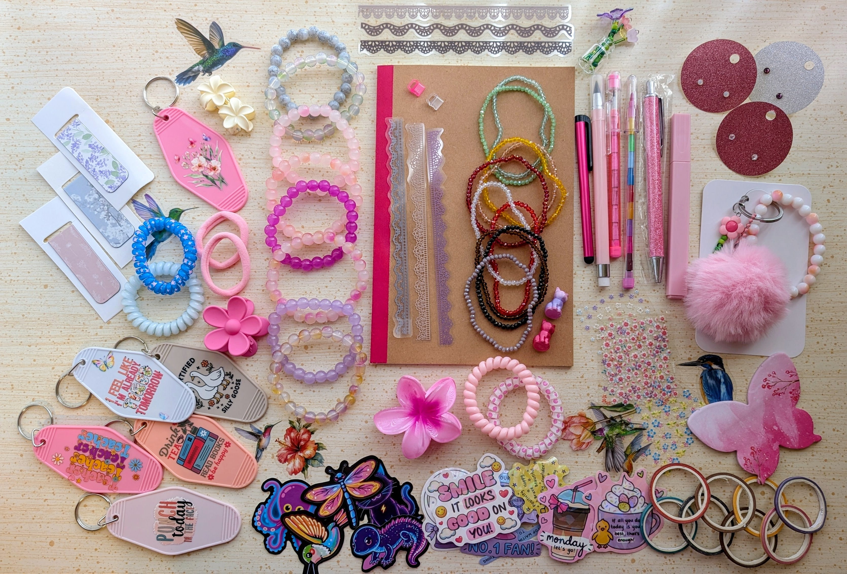 A flat-lay display of various handmade beaded bracelets, keychains, magnetic bookmarks, stickers, stationary items suck as pens, pencils, a notebook, and small craft accessories arranged on a light wood surface.