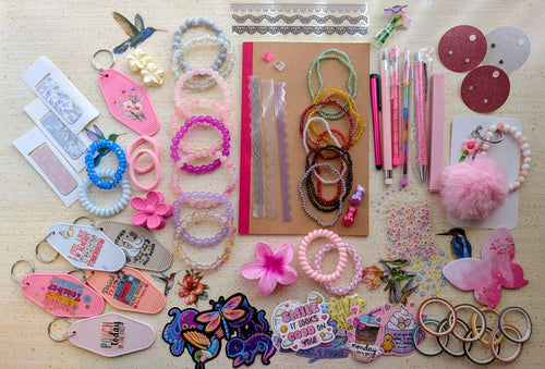 A flat-lay display of various handmade beaded bracelets, keychains, magnetic bookmarks, stickers, stationary items suck as pens, pencils, a notebook, and small craft accessories arranged on a light wood surface.