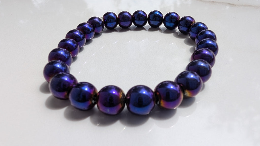 A beaded stretch bracelet made of glossy, mettalic dark purple beads with blue and violet, gold shifts, resting on a white surface.