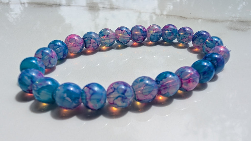A beaded stretch bracelet made of round marbled beads in shades of blue, purple, and pink, resting on a light gray marble surface.