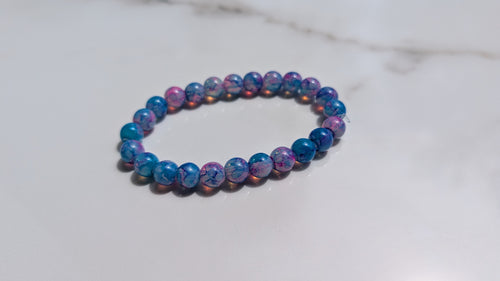 A beaded stretch bracelet made of round marbled beads in shades of blue, purple, and pink, resting on a white marble surface.