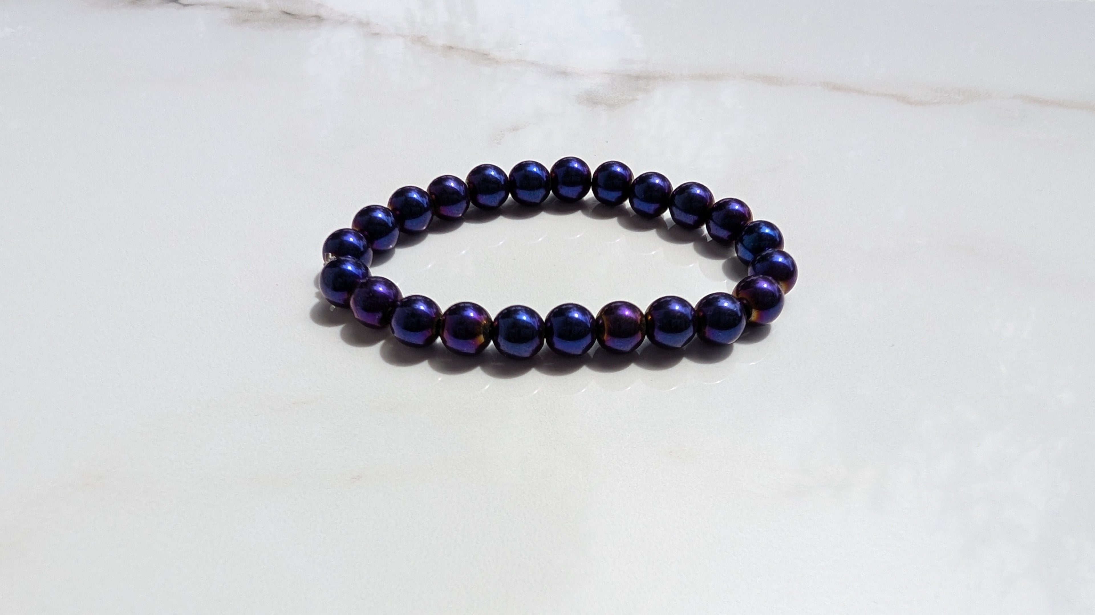 A beaded stretch bracelet made of glossy, mettalic dark purple beads with blue and violet, gold shifts, resting on a white surface.
