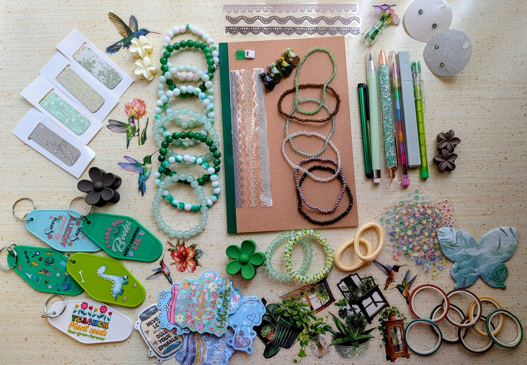 A flat-lay display of various handmade beaded bracelets, keychains, magnetic bookmarks, stickers, stationary items suck as pens, pencils, a notebook, and small craft accessories arranged on a light wood surface.