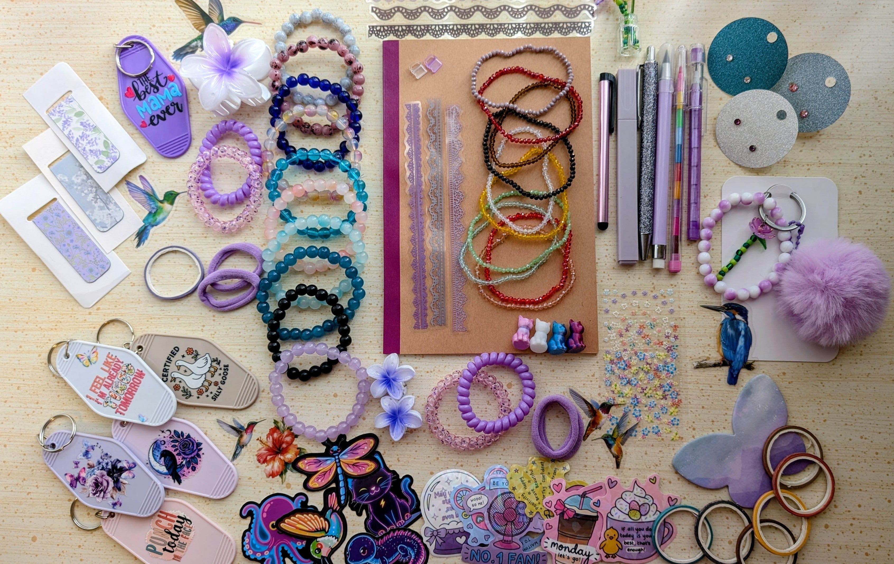 A flat-lay display of various handmade beaded bracelets, keychains, magnetic bookmarks, stickers, stationary items suck as pens, pencils, a notebook, and small craft accessories arranged on a light wood surface.