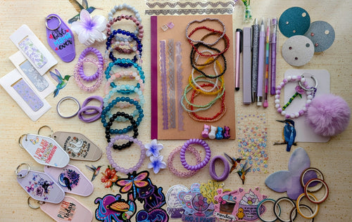 A flat-lay display of various handmade beaded bracelets, keychains, magnetic bookmarks, stickers, stationary items suck as pens, pencils, a notebook, and small craft accessories arranged on a light wood surface.