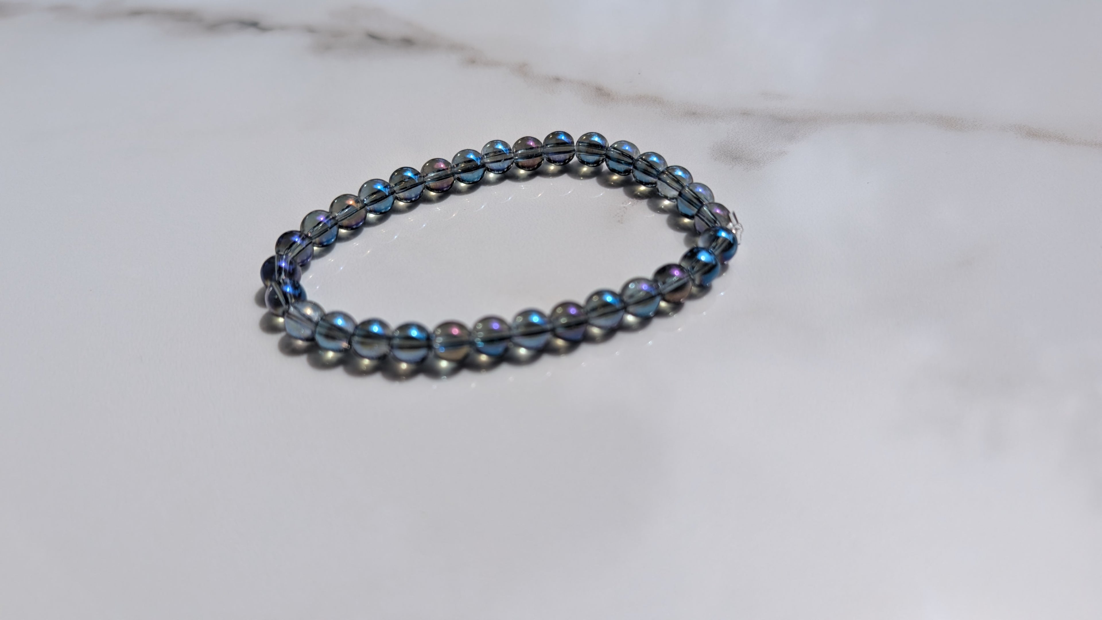 A beaded stretch bracelet with dark gray metallic beads and iridescent blue-purple accents, resting on a white marble surface.