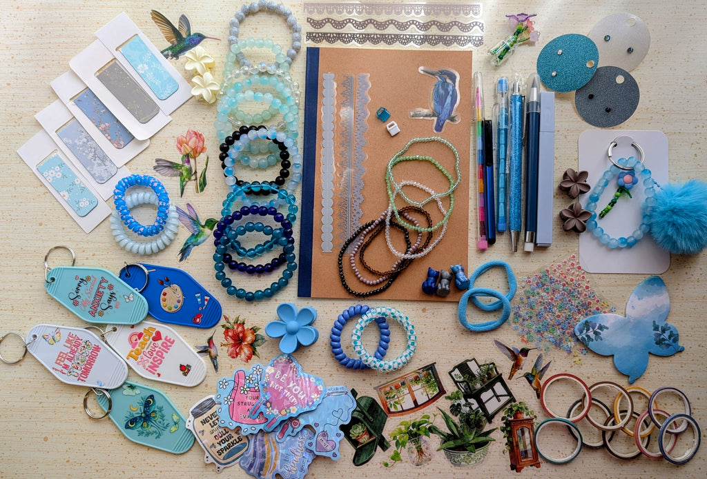 A flat-lay display of various handmade beaded bracelets, keychains, magnetic bookmarks, stickers, stationary items suck as pens, pencils, a notebook, and small craft accessories arranged on a light wood surface.