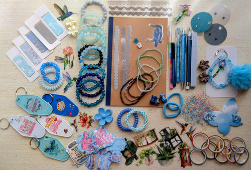 A flat-lay display of various handmade beaded bracelets, keychains, magnetic bookmarks, stickers, stationary items suck as pens, pencils, a notebook, and small craft accessories arranged on a light wood surface.